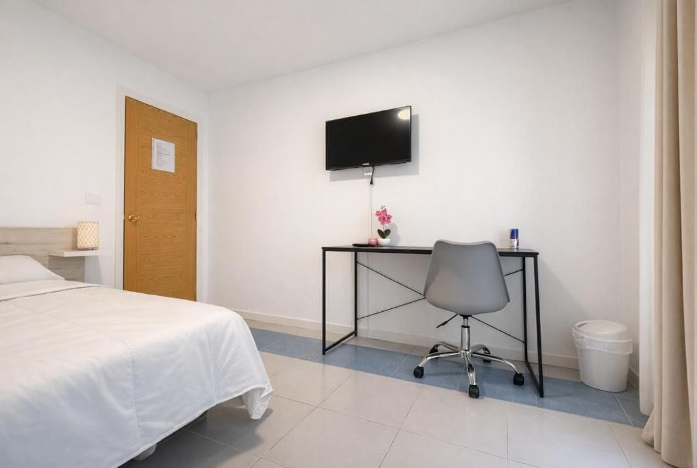 A hotel room with a bed, a desk with a chair, a television, and a trash bin.