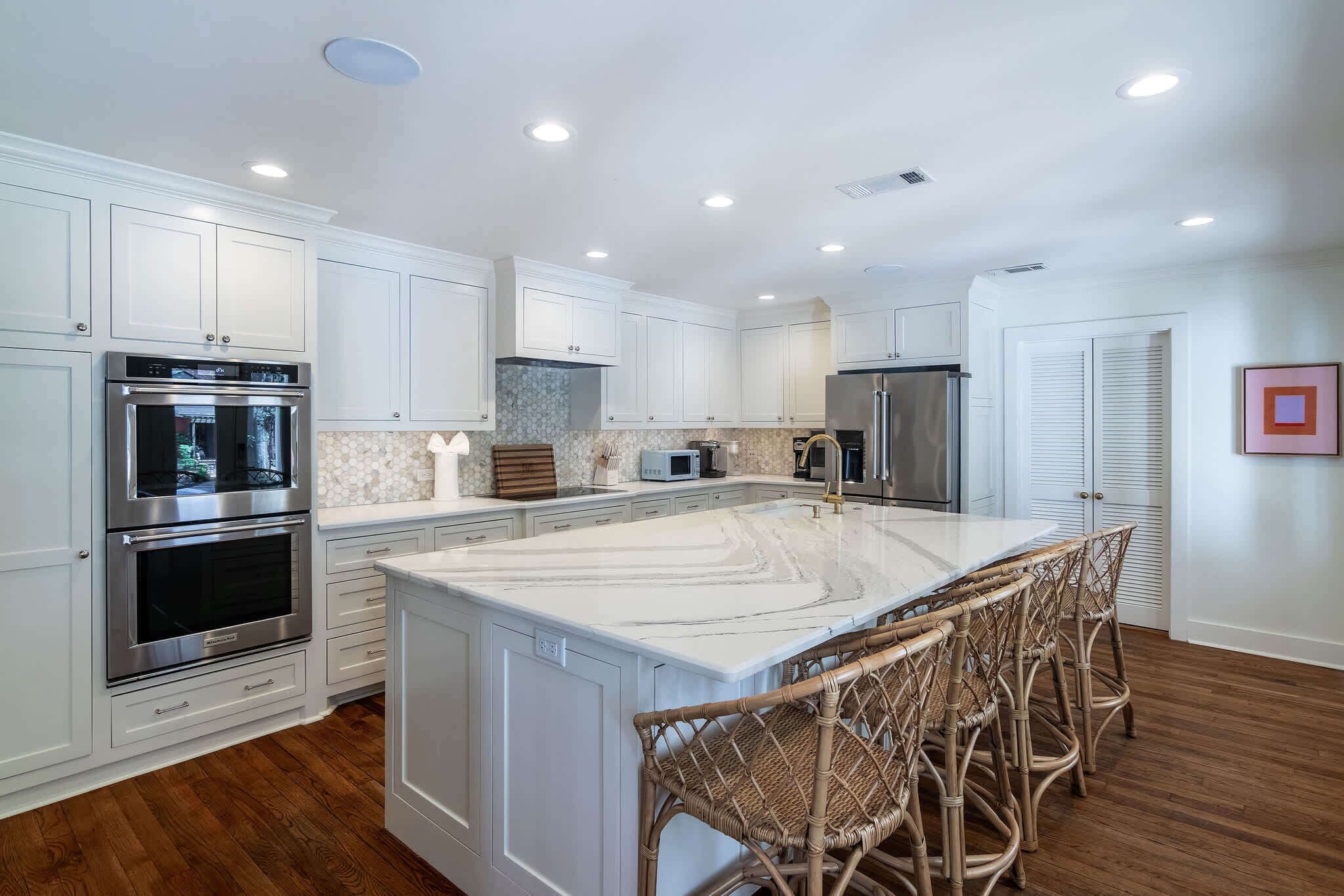A modern kitchen with white cabinetry, stainless steel appliances, a marble island, and wicker bar stools.