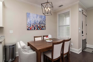 A dining area with a wooden table, four chairs, a trash can, a painting on the wall, and a window with blinds. - Hotty Toddy Hideout (Oxford)