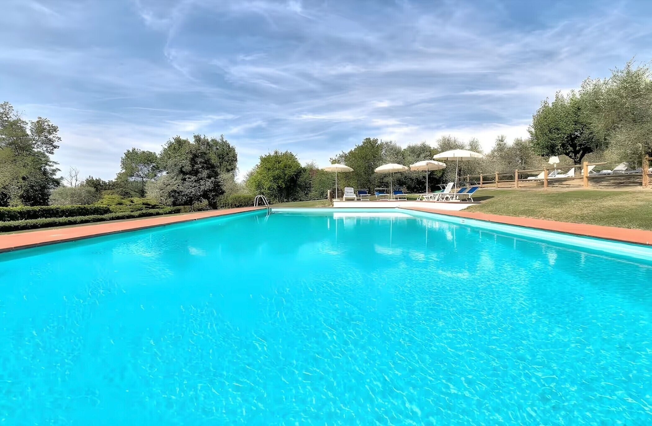 Ein klares, blaues Schwimmbecken mit Liegestühlen und Sonnenschirmen, umgeben von Bäumen und einem Holzzaun.