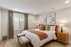 A bedroom with a bed, bedside tables, a bench, a window with curtains, and two wall-mounted lamps.