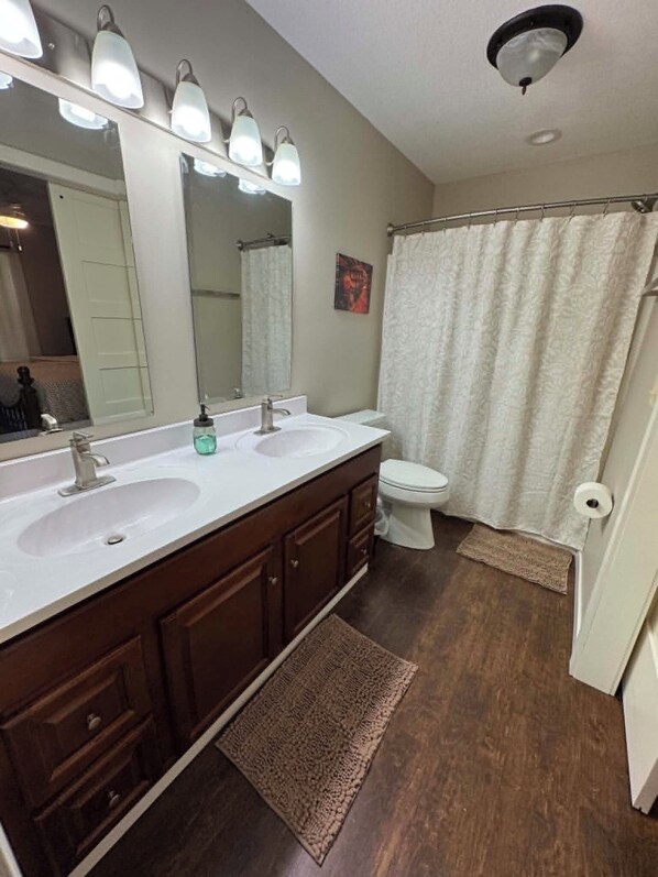 A bathroom with a double sink vanity, a toilet, a shower curtain, and a large mirror with lights. - Ken's Place (Gallipolis)