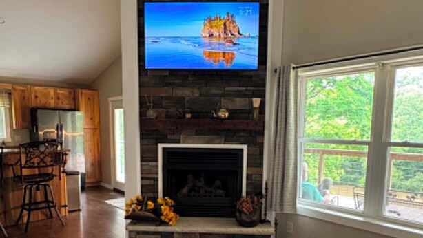 Ein Wohnzimmer mit Kamin aus Stein, einem an der Wand montierten Fernseher und einem großen Fenster.