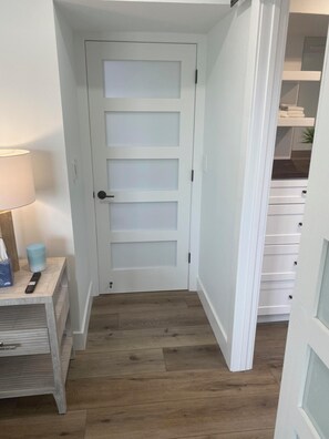 Ein schmaler Flur mit einer weißen Tür, Holzboden, einem kleinen Tisch mit Lampe und einem Schrank mit Schubladen.