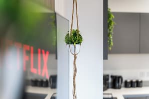 A hanging plant in a white pot suspended by a rope, with a blurred background featuring a kitchen area and a sign with red letters. - Deluxe pad– Bham Centre, Pool Table, Next to Canal +Netflix (Birmingham)