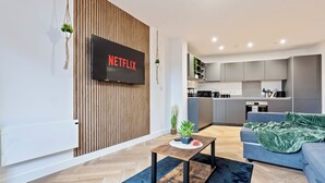 Uma sala de estar moderna com televisão de tela plana, painel de parede de madeira e uma cozinha ao fundo.