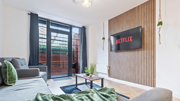 Uma sala de estar moderna com sofá, TV de tela plana, uma mesa pequena e uma porta de vidro deslizante que dá para uma área externa.