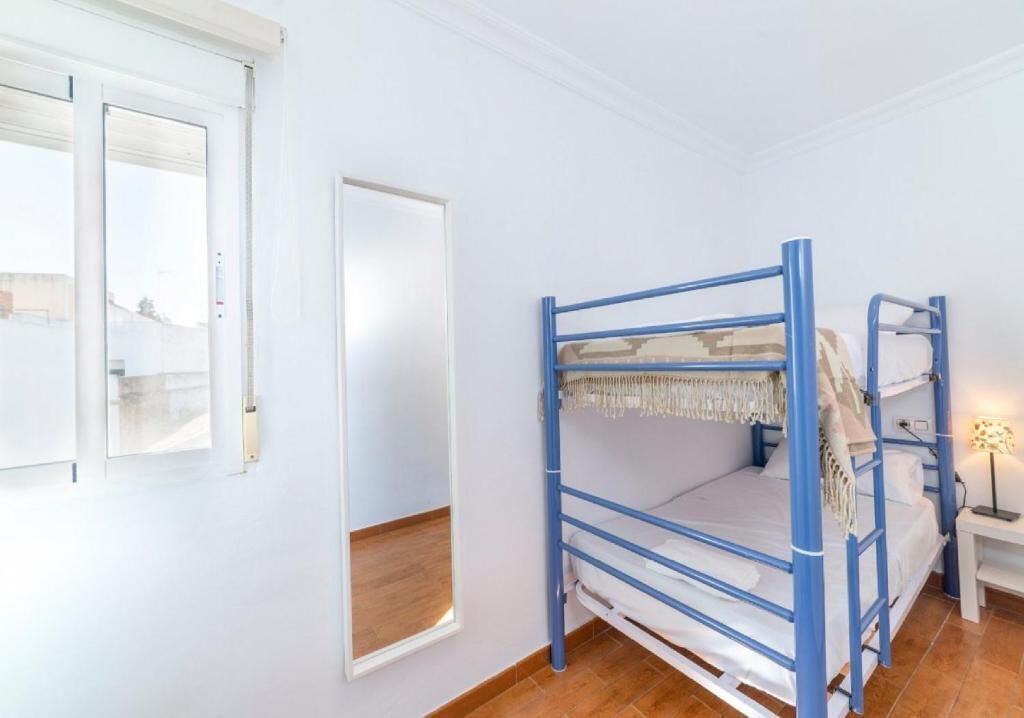 Una cama individual en literas sobre suelo de madera, pared blanca y ventana con persianas.
