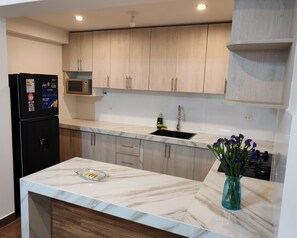 A modern kitchen with light wood cabinets, a black refrigerator, a microwave, a sink, and a marble countertop. - Apartment J&L (Cusco)