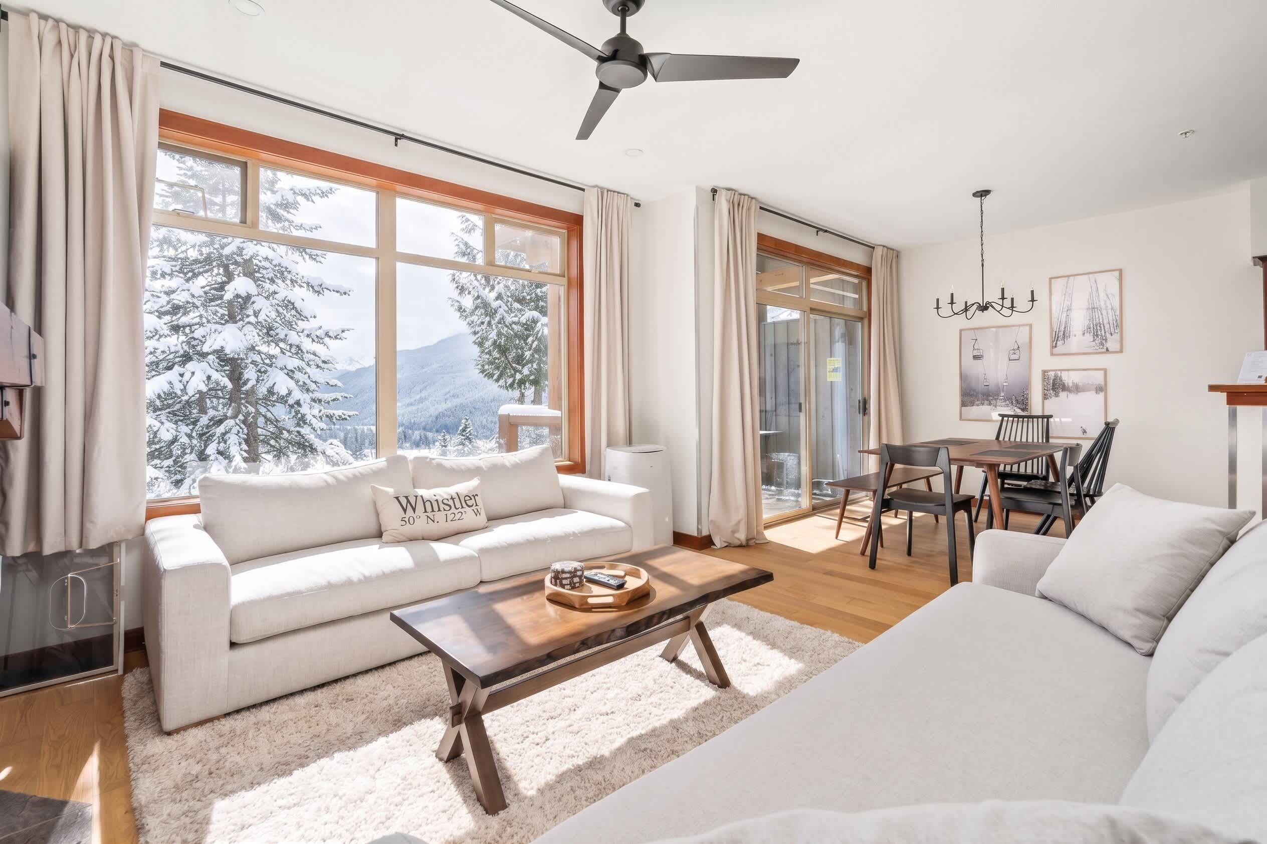 A spacious living room with a sofa, coffee table, and dining area. Large windows offer a view of snow-covered trees.