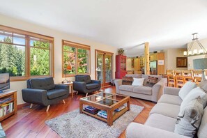 Ein geräumiges Wohnzimmer mit großen Fenstern, einem Fernseher und einem Essbereich mit Holzzmöbeln.