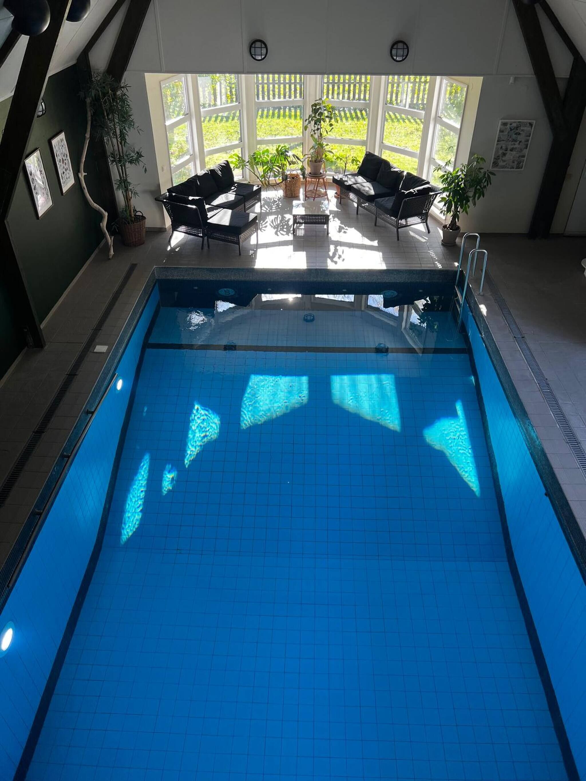 Una piscina interior con agua azul cristalina, rodeada por una zona de estar con sillones y plantas en macetas.