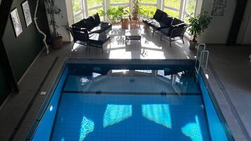 Una piscina interior con agua azul cristalina, rodeada por una zona de estar con sillones y plantas en macetas.