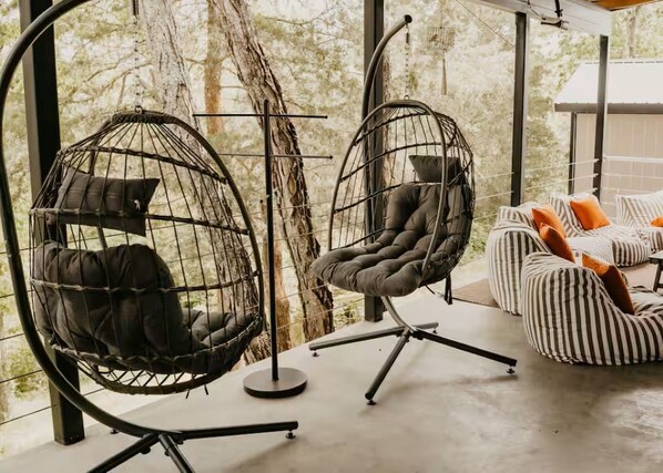 Zwei eiförmige Hängesessel mit Kissen, ein gestreiftes Sofa mit orangefarbenen Kissen und ein Betonboden.