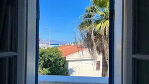 A view through an open window, revealing a building with a red-tiled roof, a palm tree, and a clear blue sky. - A Hotel  (Cesme)