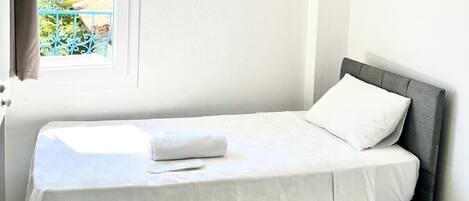 A single bed with a white mattress and pillow, a grey headboard, and a window with a view of greenery.