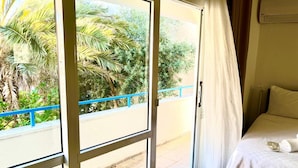 A room with a bed, a window with a view of greenery, and a balcony with a blue railing. - A Hotel  (Cesme)