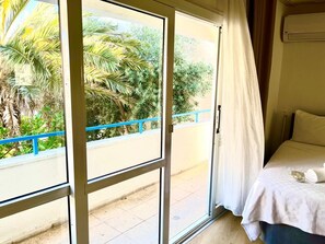 A room with a bed, a window with a view of greenery, and a balcony with a blue railing. - A Hotel  (Cesme)