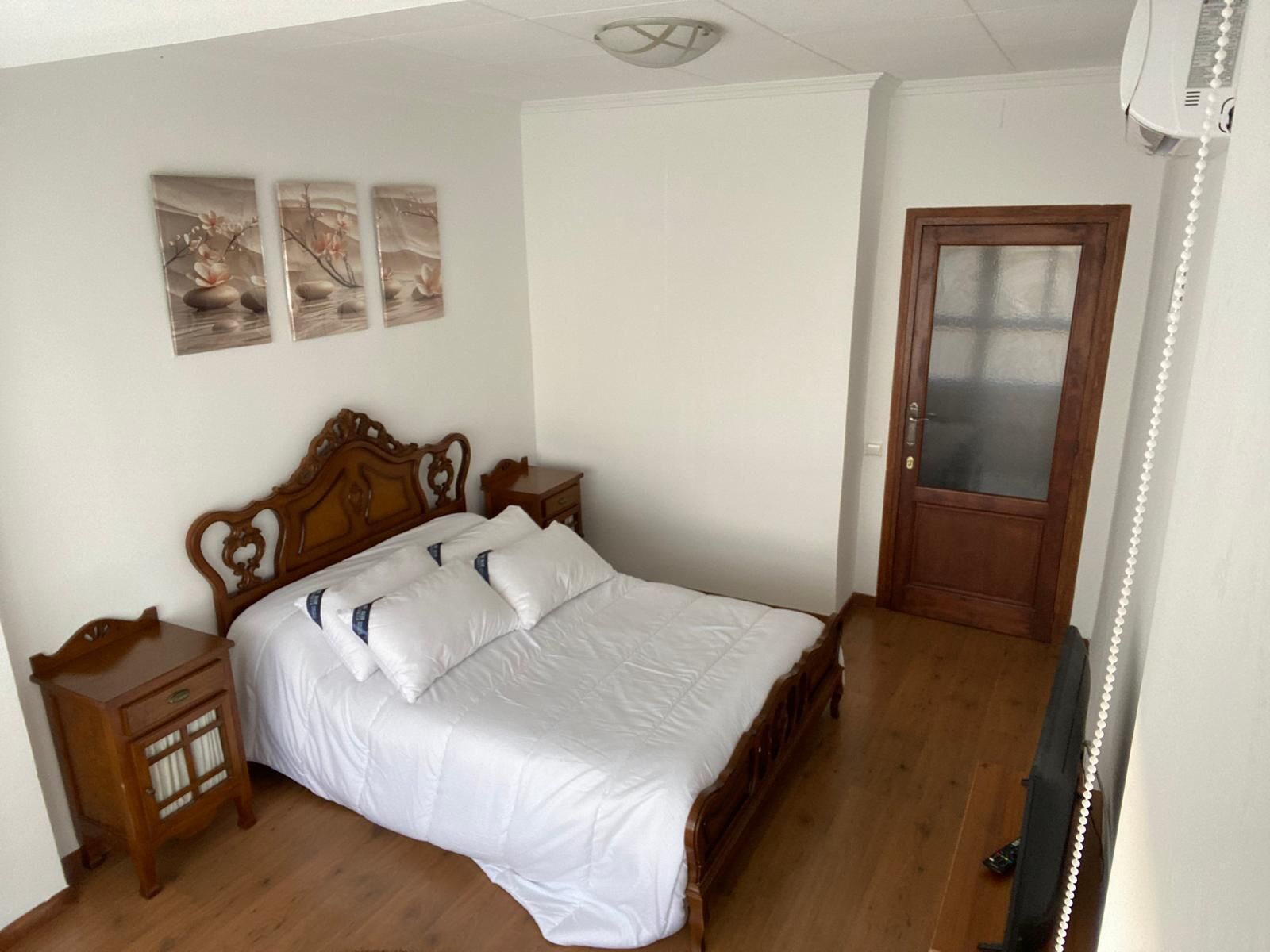 A bedroom with a wooden bed, white bedding, a wooden nightstand, a small cabinet, and a door with a frosted glass panel.