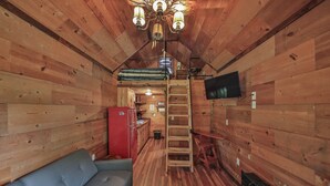 Interior acolhedor de uma cabana com sofá, televisão, escada e pequena cozinha.