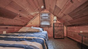 Um quarto de sótão pequeno e acolhedor, com teto inclinado, uma cama de solteiro, cômoda, abajur e janela com persianas.
