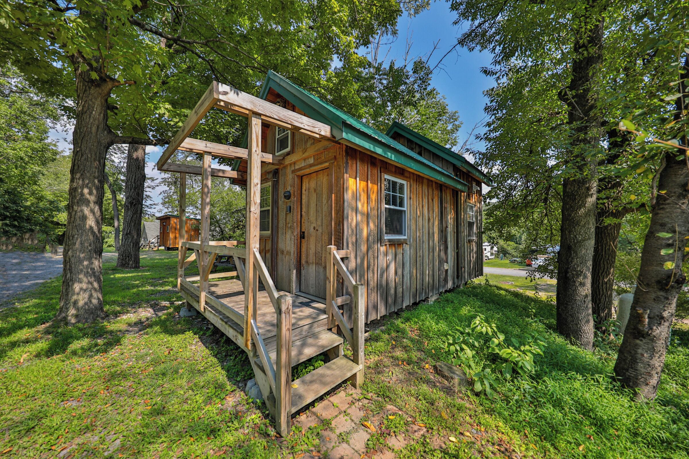 A wooden cabin with a porch and a green roof, surrounded by trees.