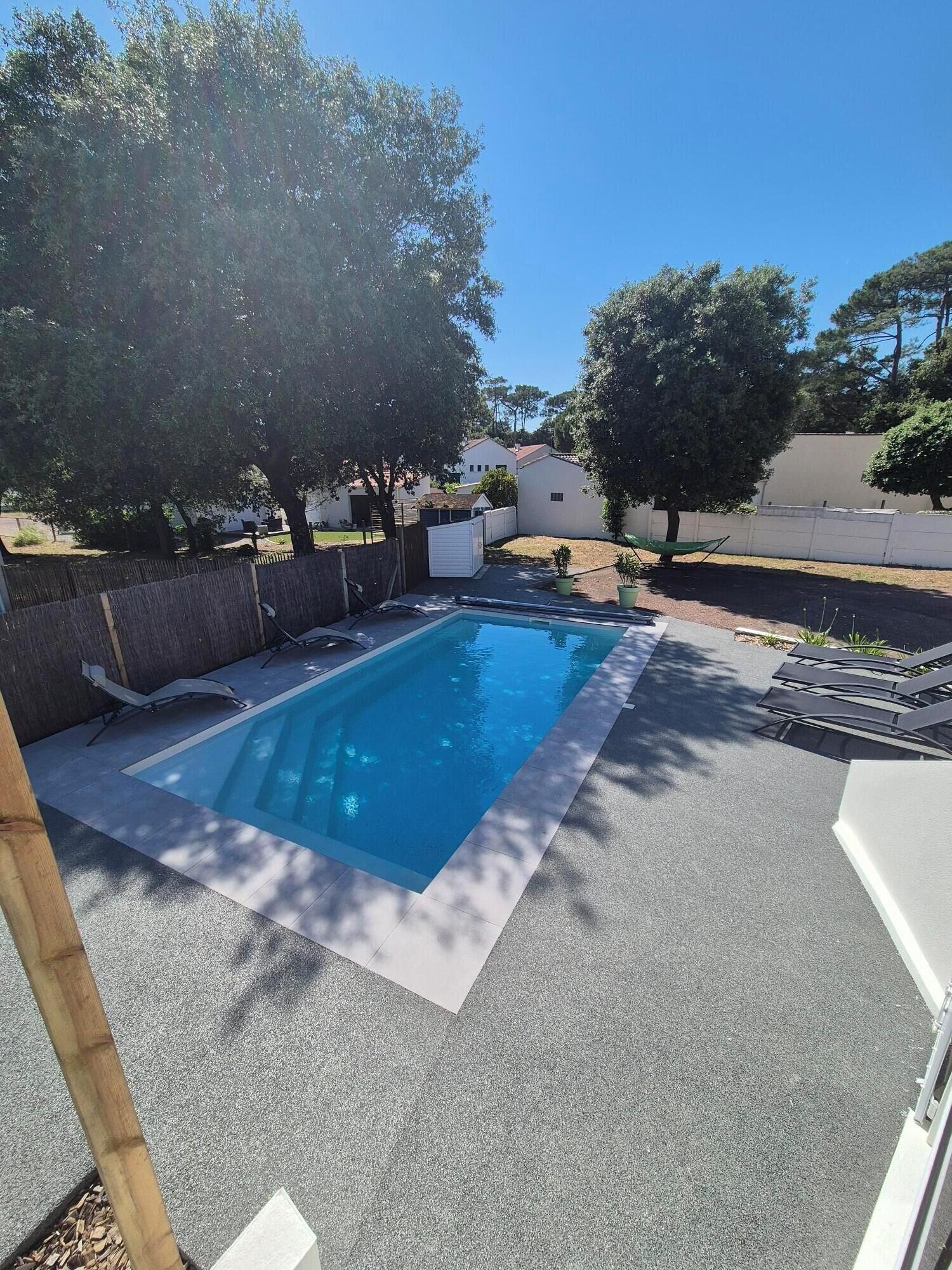 A rectangular swimming pool with a tiled deck, surrounded by trees and a white fence.