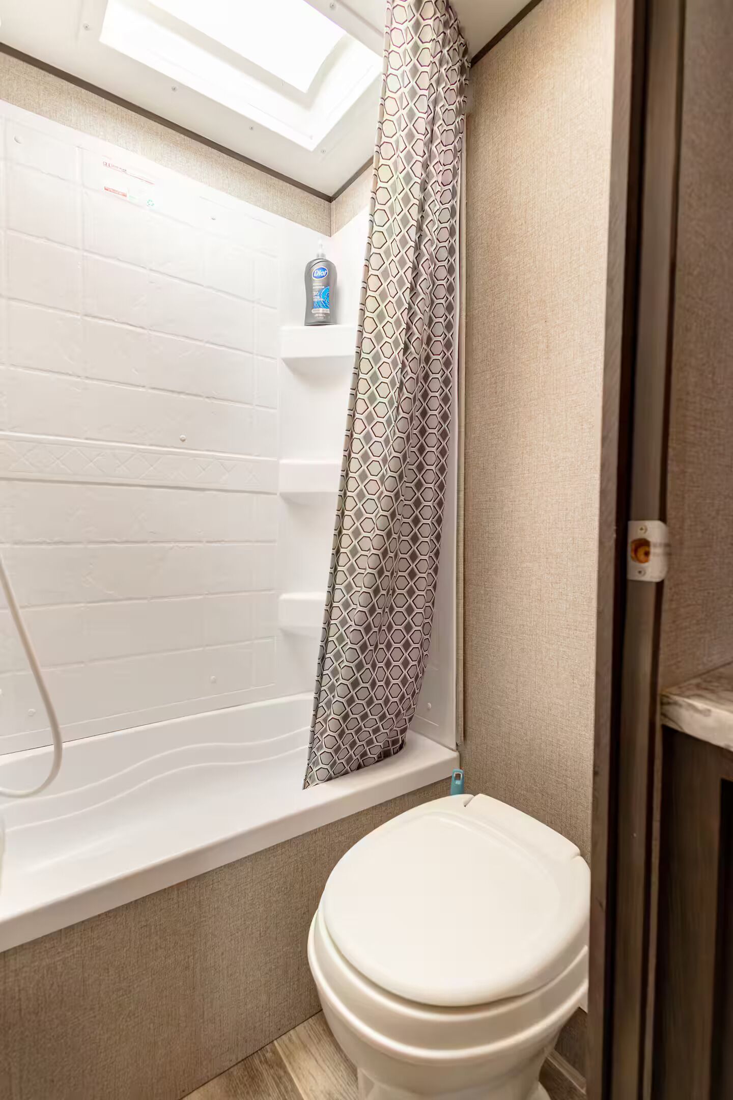 Un baño con inodoro blanco, ducha con cortina estampada y tragaluz sobre la ducha.