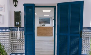 Ein Empfangsbereich mit blauen Fensterläden, einem Empfangstresen, einem an der Wand montierten Drucker und Wandfliesen mit Muster.