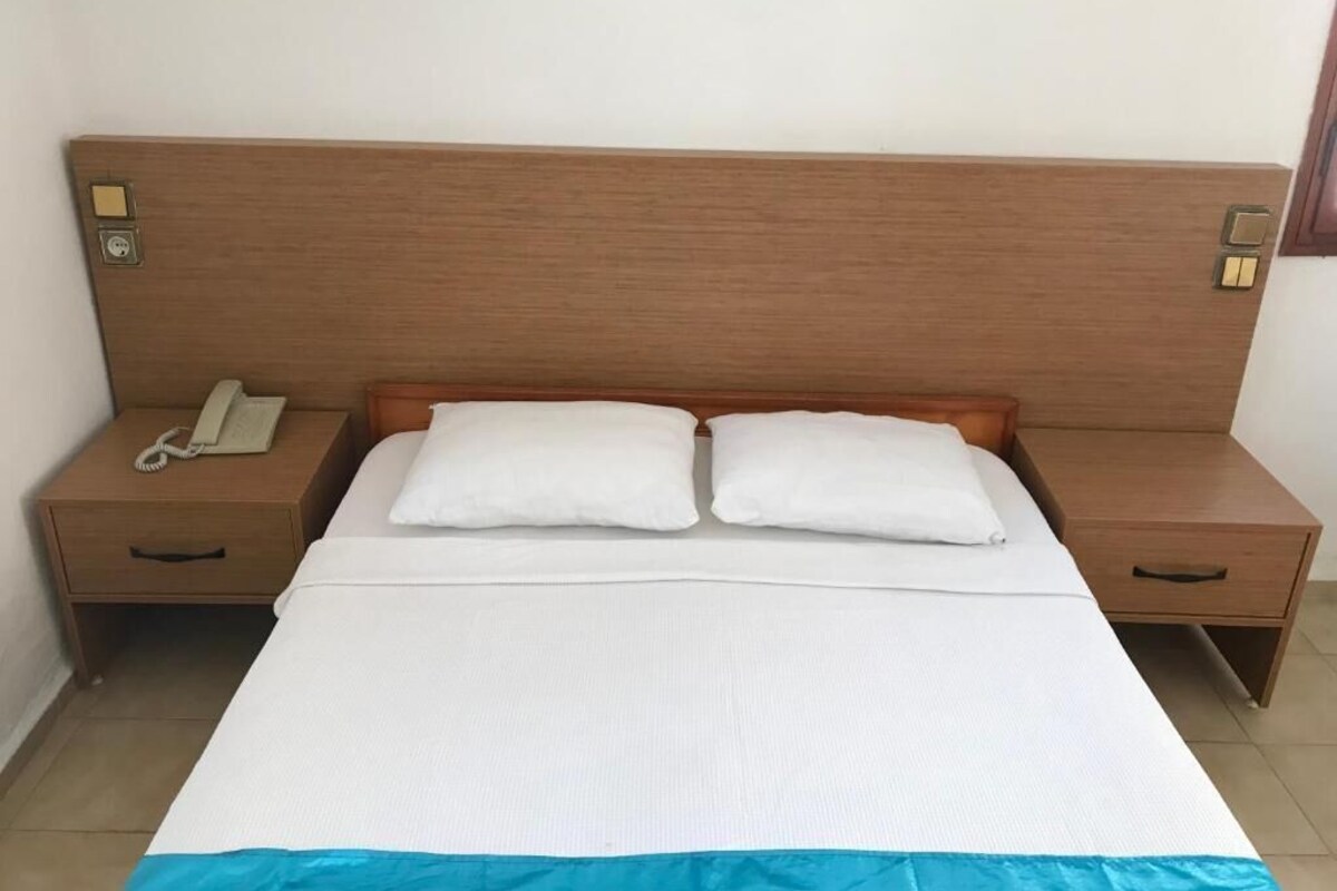 Ein Standardhotelzimmer mit einem hölzernen Kopfteil, einem Einzelbett mit weißer Bettwäsche, zwei Nachttischen und einem Telefon auf einem der Tische.