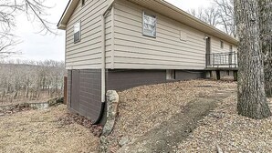 A single-story house with a dark garage door, a small porch, and a tree in the foreground.