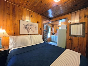 A wooden-paneled room with a bed, a painting of a horse, a mirror, and a view into the kitchen. - Willow Whisper Cottage (Hot Springs)