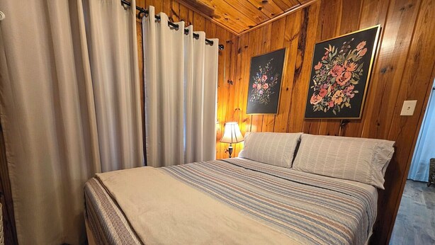 A bedroom with a bed, curtains, a lamp, and two floral paintings on the wall. - Wildflower's Escape (Hot Springs)