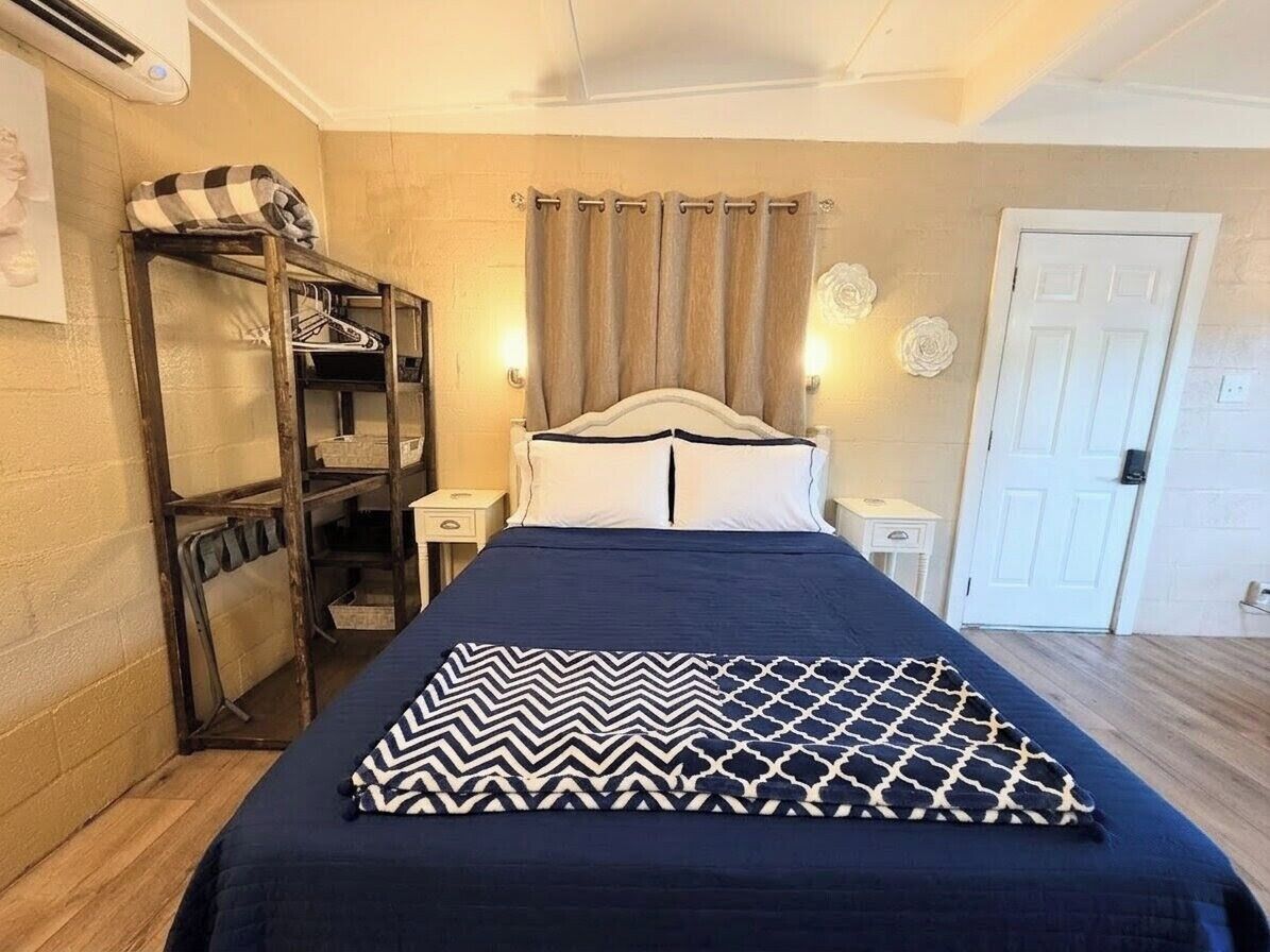 A bed with a patterned blanket, a wooden shelf with folded clothes, a nightstand with a lamp, and a door.