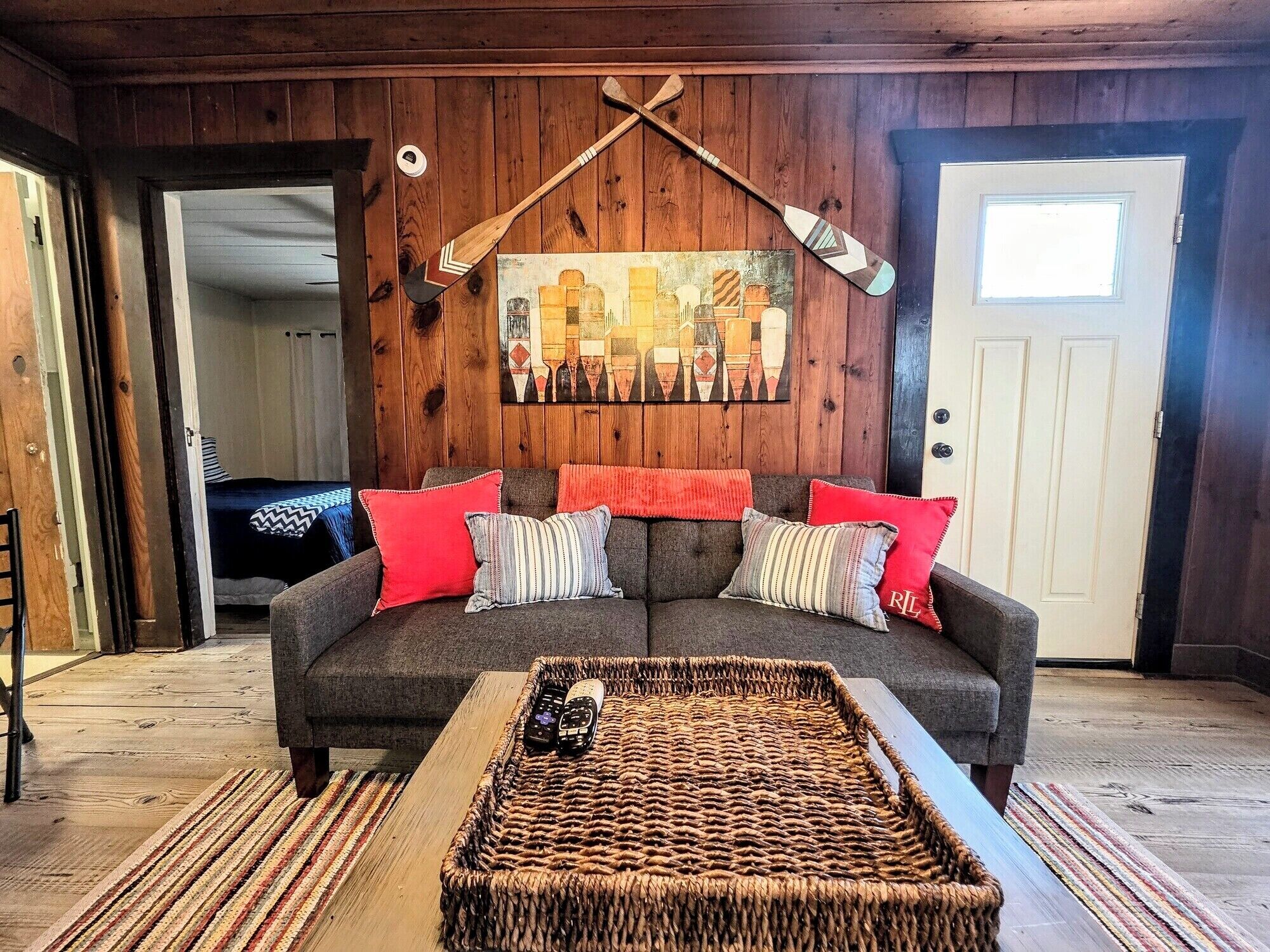 A cozy living room with a grey sofa, red and striped pillows, a woven coffee table, and a cityscape painting on the wall.
