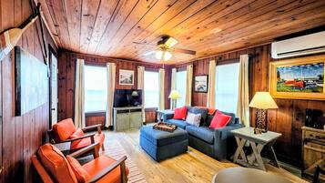 A cozy cabin interior with wooden walls, a ceiling fan, a sofa set, a coffee table, and a television.