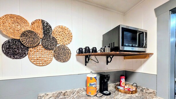 A kitchen with a granite countertop, a microwave, a shelf with mugs, and a basket of bread. - Pebble Beach Cottage (Hot Springs)