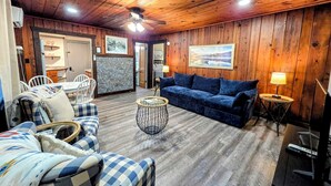 A cozy room with a wooden ceiling, a blue sofa, a checkered couch, a round coffee table, and a television. - Pebble Beach Cottage (Hot Springs)