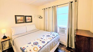 A bedroom with a bed, a nightstand, a ceiling fan, a window with curtains, and a wall-mounted air conditioning unit. - Serenity Shores Cottage (Hot Springs)