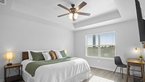 A bedroom with a bed, a desk, a chair, a ceiling fan, a window, and a nightstand with a lamp. - Brand-New Med District Home (Temple)
