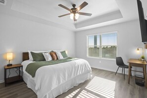 Ein Schlafzimmer mit Bett, Schreibtisch, Stuhl, Deckenventilator, Fenster und Nachttisch mit Lampe.