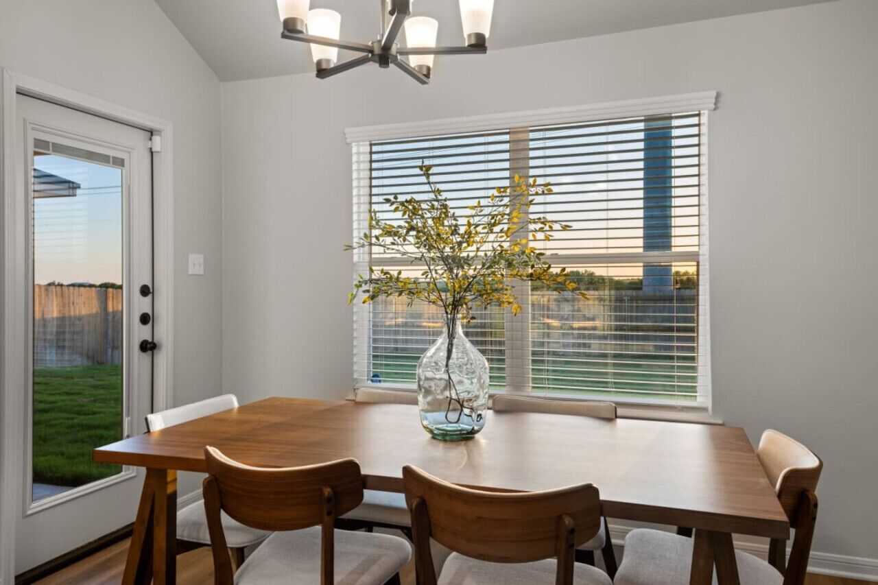 A dining room with a wooden table, chairs, a vase with flowers, a window with blinds, and a door.
