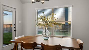 A dining room with a wooden table, chairs, a vase with flowers, a window with blinds, and a door.