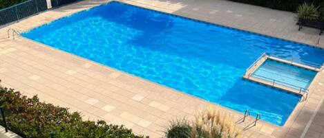 A rectangular swimming pool with a tiled deck, surrounded by greenery and residential buildings.