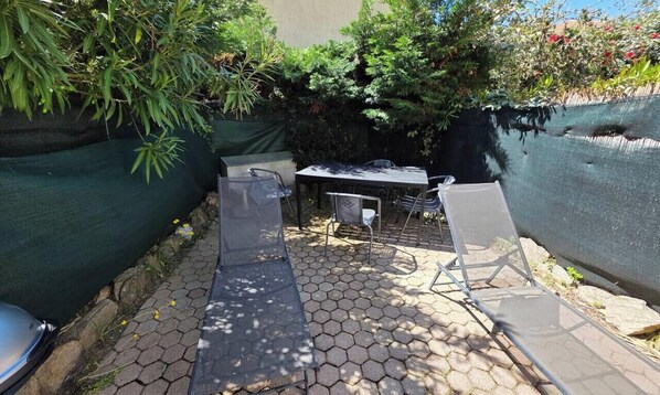 Un patio con una mesa y sillas, rodeado de plantas y una cerca verde.