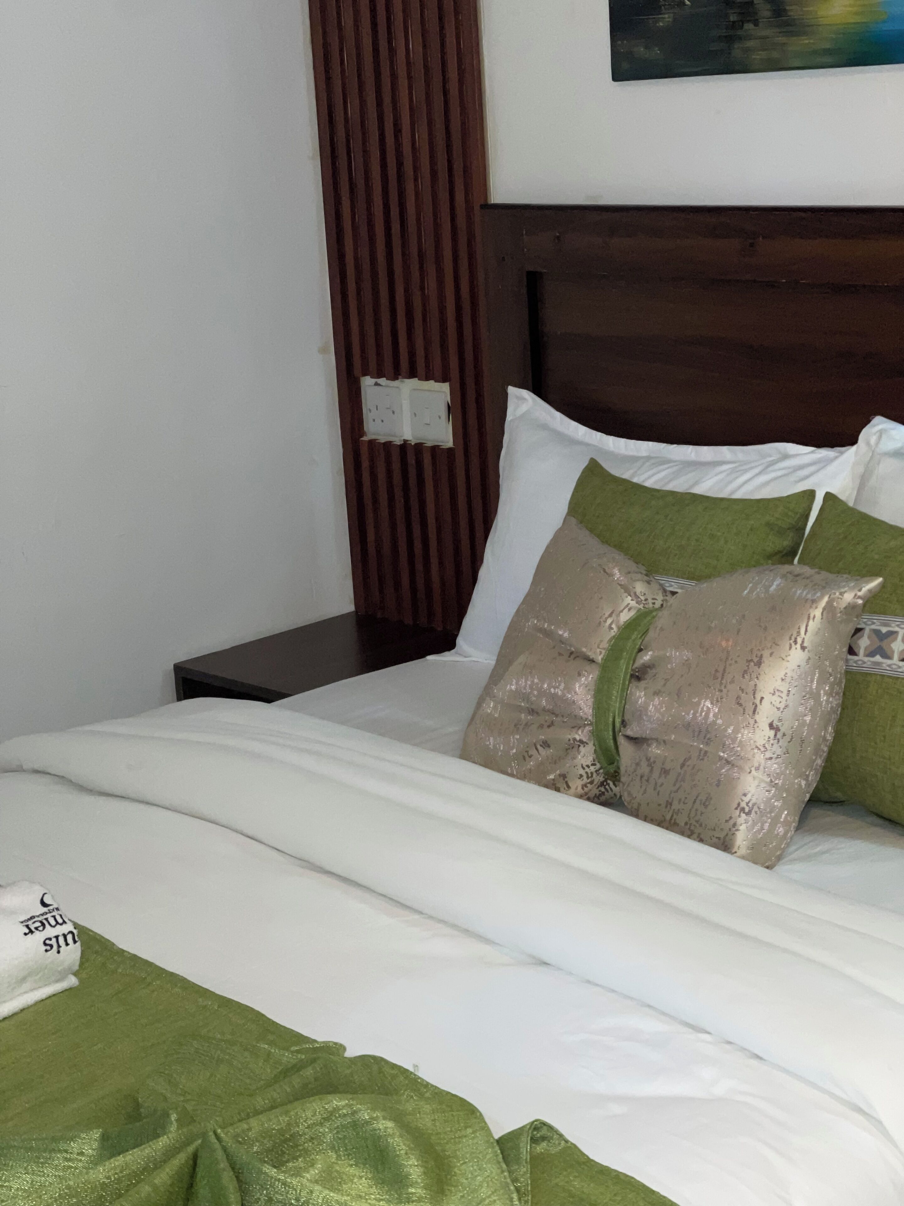 Een net opgemaakt bed met wit en groen beddengoed, een houten hoofdbord en een klein nachtkastje.