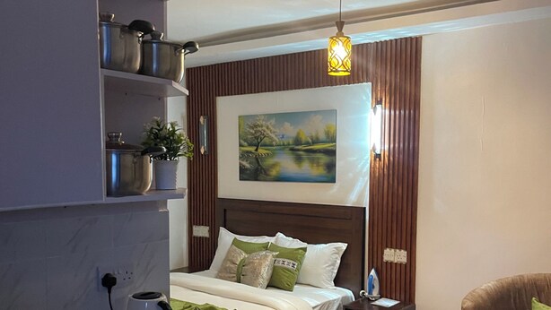 A compact hotel room with a kitchenette, a bed with green and white bedding, a painting of a landscape, a round table with decorative items, and a brown armchair.