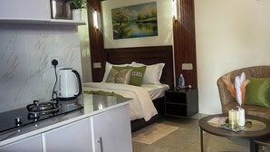 A compact hotel room with a kitchenette, a bed with a headboard, a small round table with a vase, and a painting on the wall.