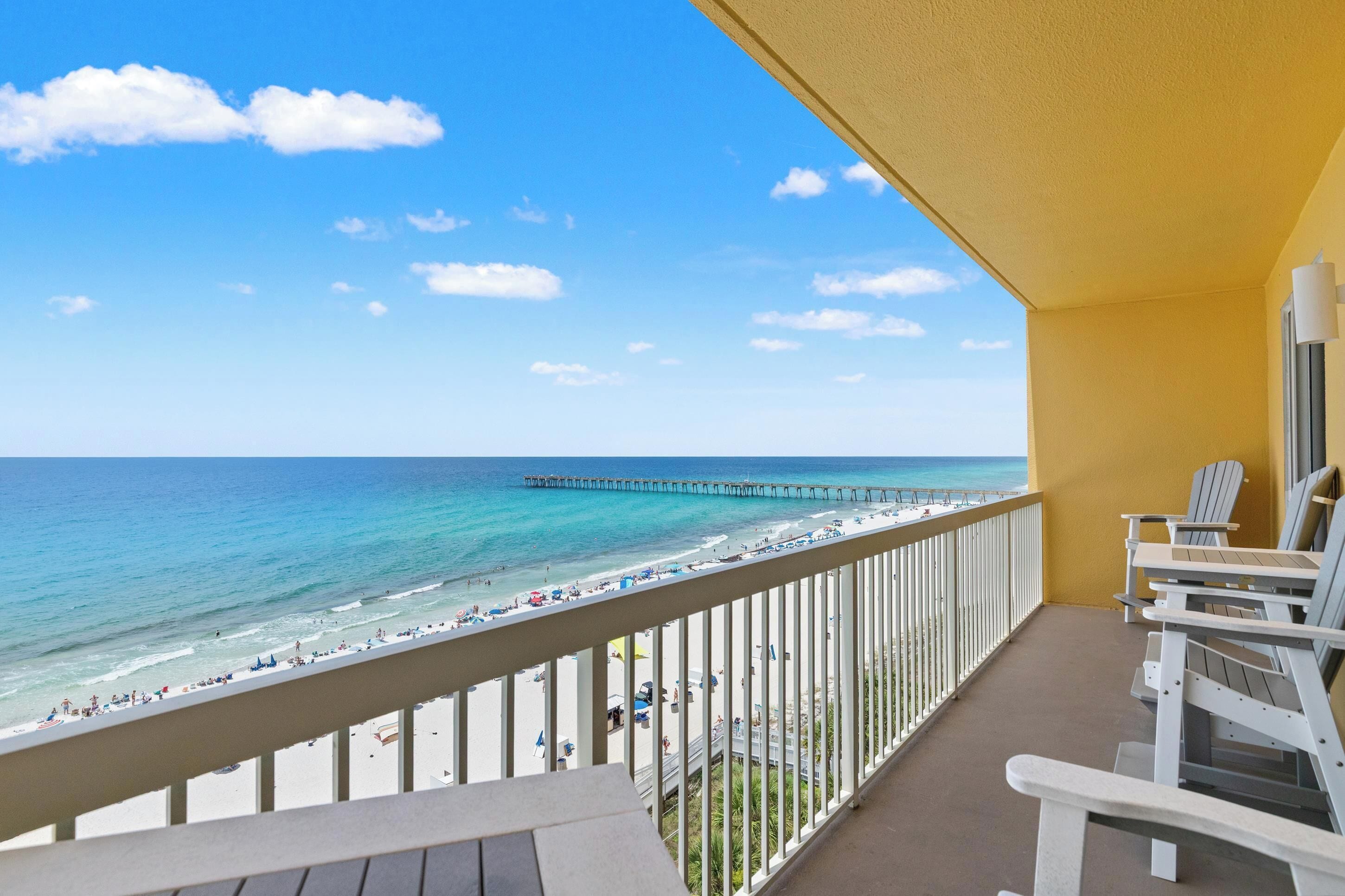 A balcony with a view of the beach and ocean, white chairs, and a wooden deck.