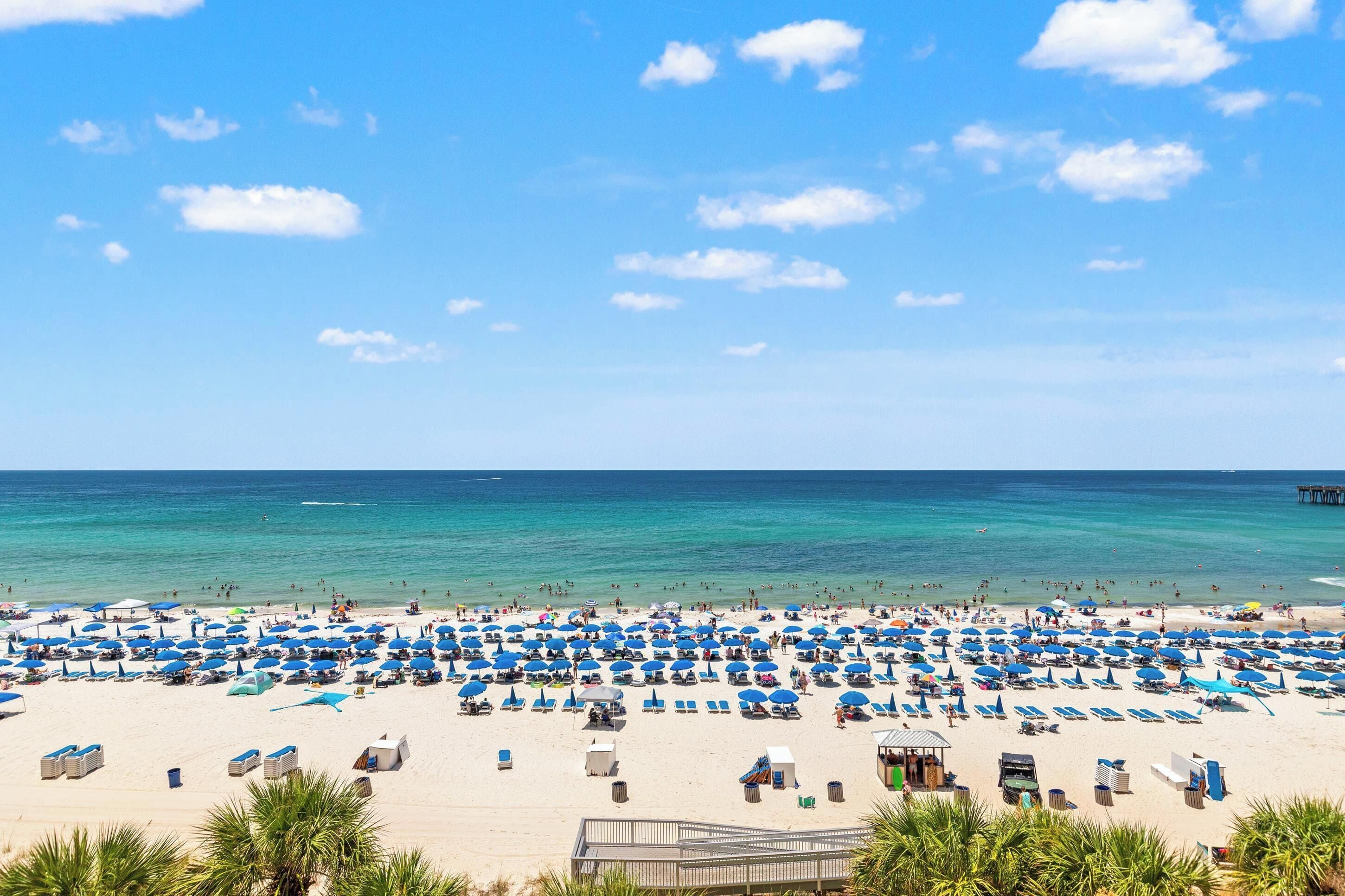 Una playa con numerosas sombrillas azules y sillas de descanso, un muelle de madera a lo lejos y cielos azules despejados.